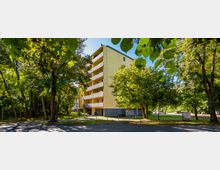 Mehrfamilienhaus mit gelber Fassade und mehreren Balkonen, umgeben von Bäumen und Grünflächen; im Vordergrund verläuft eine Straße, während im Hintergrund weitere parkende Autos sichtbar sind.