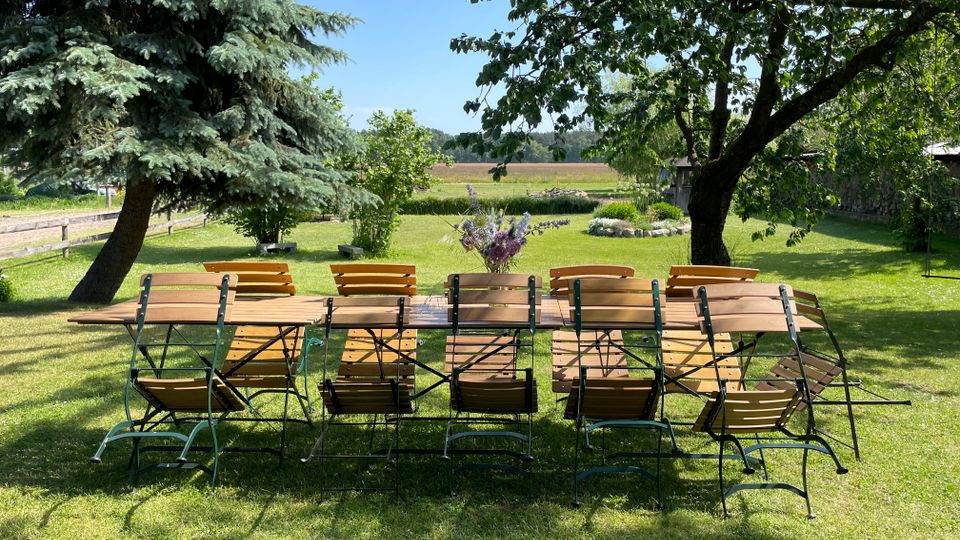 Langer Holzgartentisch mit mehreren Klappstühlen auf einer gepflegten Rasenfläche unter großen Bäumen. Im Hintergrund sind ein ländlicher Garten mit Sträuchern und Wiesen sowie Felder am Horizont zu sehen.