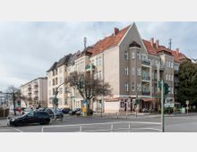 Straßenecke mit mehrstöckigen Wohnhäusern im Altbaustil, gekennzeichnet durch helle Fassaden, rote Ziegeldächer und Balkone. Im Erdgeschoss befinden sich Geschäfte und Cafés, und der Bereich ist von parkenden Autos, Fußgängern und Ampeln an der Kreuzung geprägt.