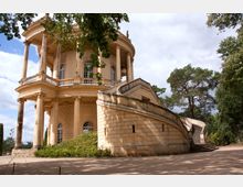 Runder, klassizistischer Pavillon mit Säulengang und einer geschwungenen Treppe aus Sandstein, umgeben von Bäumen und einem sandigen Platz. Im Hintergrund sind vereinzelte Kiefern und ein bewölkter Himmel zu sehen.