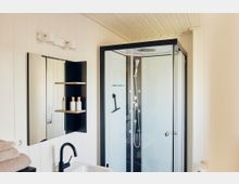 Modernes Badezimmer mit heller Holzwandverkleidung, einer Glas-Duschkabine mit schwarzen Rahmen, einem Waschbecken mit schwarzem Wasserhahn und einem Wandspiegel mit integriertem Regal für Pflegeprodukte.