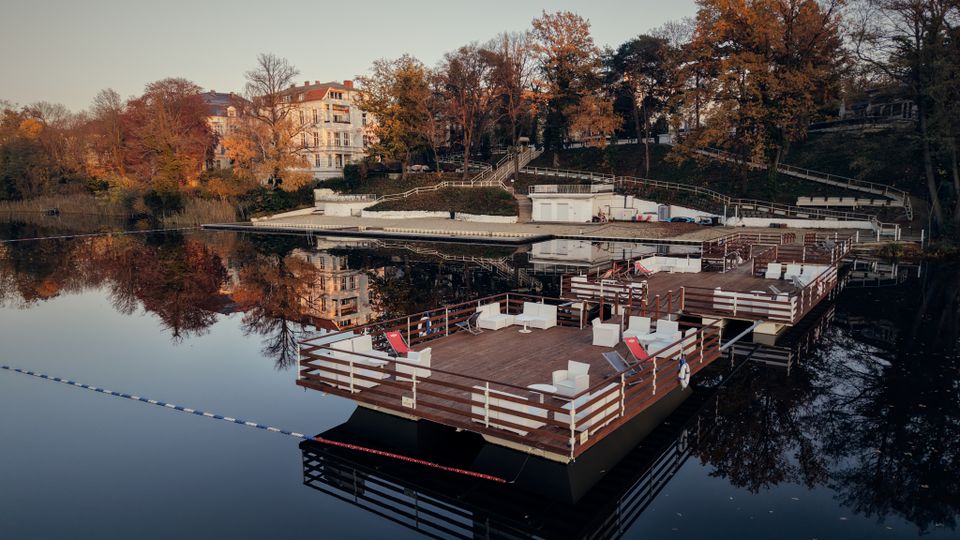 A serene waterfront scene with wooden floating platforms furnished with chairs and tables, surrounded by calm reflective water. In the background, there are trees in autumn colors, a hillside with pathways, and several white multi-story buildings nestled among the foliage.
