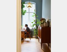 Arbeitszimmer mit Pflanzen und Holzmöbeln. Ein Mensch sitzt an einem Schreibtisch vor einem großen Fenster mit Rundbogen und hellem Tageslicht, umgeben von Zimmerpflanzen; rechts steht eine retro-inspirierte Holzkommode.