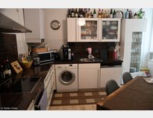 Moderne, kompakte Küche mit weißen Schränken, dunklen Arbeitsplatten und Fliesenspiegel; ausgestattet mit Elektrogeräten wie Waschmaschine, Kaffeemaschine, Mikrowelle und Herd. Im Vordergrund ein Esstisch, an der Wand eine Uhr, und Gläser sowie Flaschen sind in einem Schrank und auf Oberschränken verstaut.