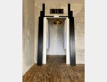 Ein schmaler Raum mit einem Türdurchgang, umrahmt von schwarzen vertikalen und horizontalen Elementen, vor einer Betonwand. Der Boden besteht aus rohem Holz, und eine goldene Deckenleuchte ist im angrenzenden Raum sichtbar.