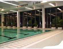 Hallenbad mit beleuchtetem Schwimmbecken, gefliestem Boden und großen Glasfenstern im Hintergrund. Entlang der Fensterreihe stehen Pflanzen in großen, weißen Blumentöpfen. Ein Schild zeigt die Wassertiefe von 2,20 m an.