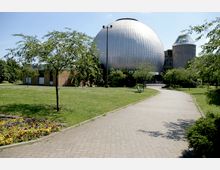 Ein moderner Gebäudekomplex mit einer großen, kugelförmigen, silbernen Konstruktion in der Mitte, umgeben von Rasenflächen, kleinen Bäumen und Blumenbeeten; ein gepflasterter Weg führt durch die Grünanlage.