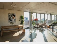 Heller, moderner Innenraum mit Holzmöbeln, großen Fensterfronten und Blick auf eine Außenterrasse. An der Wand hängt ein abstraktes Kunstwerk, während Pflanzen und dekorative Elemente den Raum ergänzen.