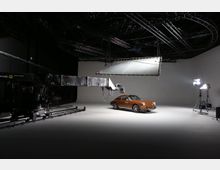 Ein Fotostudio mit einem braunen Oldtimer-Auto im Mittelpunkt, umgeben von professionellem Beleuchtungsequipment, einem Kamerakran auf Schienen und einer großen Hängeleuchte. Der Raum hat einen neutralen weißen Hintergrund und eine teilweise sichtbare schwarze Deckenstruktur.