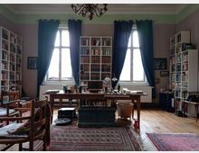 Ein gemütliches Arbeitszimmer mit einem großen Holztisch in der Mitte, umgeben von hohen Bücherregalen, die mit Büchern gefüllt sind. Zwei große Fenster mit dunklen Vorhängen lassen Tageslicht einfallen; der Boden ist aus Holz und teilweise mit Teppichen bedeckt.