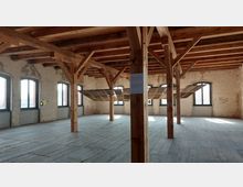 Innenraum eines historischen Gebäudes mit Holzbalkenkonstruktion und Holzboden. Die Wände sind aus unverputztem Mauerwerk, und mehrere große Fenster mit Rundbögen lassen Tageslicht herein; in der Mitte ist eine hängende Holzkonstruktion erkennbar.