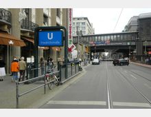 Straßenszene an der Friedrichstraße in Berlin mit einem blauen U-Bahn-Schild, das den U-Bahnhof Friedrichstraße anzeigt, mehreren Geschäften mit Markisen auf der linken Seite und dem Bahnhof Friedrichstraße mit einem Stahl-Glas-Gebäude über der Straße im Hintergrund. Straßenbahngleise und Fahrzeuge sind auf der Straße zu sehen, während Passanten den Gehweg nutzen.