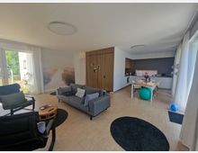 Wohnzimmer mit offener Küche in modernem Stil. Der Raum enthält eine graue Couch, schwarze Sessel, einen Holztisch mit weißen Stühlen, eine Kochecke mit weißen Fronten und Holzdetails, sowie große Fenster mit Blick auf einen Garten.