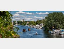 Flusslandschaft mit mehreren Booten auf dem Wasser, daneben eine Uferpromenade mit kleinen Gebäuden und Bäumen; im Hintergrund ist ein Kirchturm zwischen grünem Laub und Wolkenhimmel sichtbar.