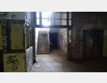 Verfallenes Treppenhaus in einem verlassenen Gebäude mit vergilbten Wänden und zahlreichen Graffiti; ein schmaler Zugang führt zu einer Treppe mit Geländern, die nach oben führt, während der Raum umgeben ist von bröckelnder Farbe und herunterhängenden Kabeln.