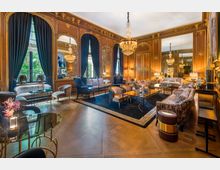 Luxuriös eingerichteter Salon mit hölzernen Wänden und detaillierten goldenen Verzierungen, großen Spiegeln und großzügigen Fensterfronten mit schweren dunkelblauen Vorhängen. Der Raum ist mit eleganten Sofas, Sesseln, Glastischen und Kristallleuchtern ausgestattet, die eine raffinierte Atmosphäre schaffen.