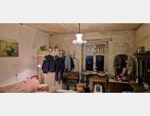 Ein einfach eingerichtetes Zimmer mit abgenutzten Wänden und Decke, einem Sofa, einem Schrank mit daran aufgehängter Kleidung und unterschiedlichen Möbelstücken. Im Hintergrund ein altes Fenster mit Vorhängen, flankiert von einem ovalen Spiegel auf einer Kommode.