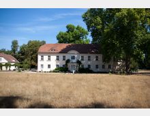 Historisches Herrenhaus mit heller Fassade und rotem Satteldach, umgeben von einem trockenen Grasfeld und großen Bäumen im Hintergrund. Links daneben steht ein Nebengebäude mit Dachbegrünung.