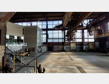 Innenansicht einer großen Industriehalle mit hohen, verglasten Fenstern und sichtbarer Stahlkonstruktion. Im Vordergrund eine moderne Ausstellung mit Informationstafeln, Treppen und historischen Exponaten; an der Decke sind alte Kräne und Maschinengerüste zu sehen.