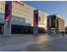 Außenbereich eines modernen Gebäudekomplexes mit einer beschrifteten Fassade „Music Hall“. Im Vordergrund befinden sich mehrere große, graue Begrenzungssäulen mit digitalen Werbedisplays. Der Boden ist mit hellgrauen Platten gepflastert, und rechts im Bild ist ein weiteres Gebäude mit zahlreichen Fenstern zu sehen.