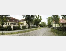 Straßenszene mit historischen Gebäuden: Links ist ein gelbes Gebäude mit rotem Ziegeldach und kleinen Fenstern, umgeben von einem Zaun und Bäumen. Rechts führt eine schmale, gepflasterte Straße vorbei an weiteren Gebäuden und Grünflächen.