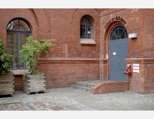 Backsteinfassade eines historischen Gebäudes mit zwei großen Rundbogenfenstern und einem Eingang mit grauer Doppeltür, über der „Aufgang zur Bücherei“ steht. Links und rechts des Eingangs befinden sich Pflanzkästen aus Holz sowie ein Hydrant an der rechten Gebäudeecke. Pflastersteine und Beton bilden den Bodenbelag.