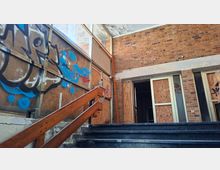 Verlassener Eingangsbereich eines Gebäudes mit Backsteinwänden, beschädigter Decke und einem Treppenaufgang mit Holzgeländer. Große Fensterflächen und Türen sind mit Sperrholz vernagelt und mit Graffiti besprüht.