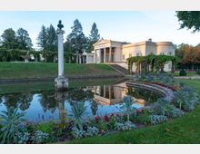 Klassizistisches Gebäude mit Säulenfassade, vor einem symmetrischen Teich mit Blumenbeeten und einer zentralen Marmorsäule. Im Hintergrund befinden sich Pergolen mit grünem Bewuchs und eine Rasenfläche, umgeben von Bäumen.