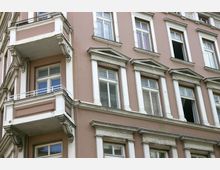 Ansicht einer historischen Fassade eines Wohngebäudes mit rosafarbenem Putz und weißen Fensterrahmen. Die Architektur ist durch Zierleisten, dekorative Giebel über den Fenstern und einen kleinen offenen Balkon mit verzierten Konsolen geprägt.