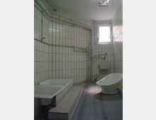 Ein einfach gefliestes Badezimmer mit weißen Wandfliesen, sichtbaren Rohren, einer erhöhten Badewanne, einem kleinen Waschbecken, einer freistehenden Schüssel sowie einem ablaufgitterartigen Boden. Ein kleines Fenster mit Gitter sorgt für begrenzten Lichteinfall.