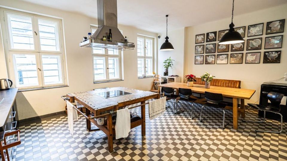 Helle Wohnküche mit großen Sprossenfenstern und schwarz-weiß kariertem Fliesenboden, in der Mitte eine Kochinsel aus Holz mit integrierter Herdplatte und markanter Dunstabzugshaube. Rechts steht ein langer Holztisch mit Stühlen und gepolsterter Bank unter schwarzen Pendelleuchten, an der Wand eine Reihe gerahmter Bilder.