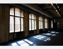 Innenraum einer ehemaligen Industriehalle mit hohen Decken und großen, bogenförmigen Sprossenfenstern entlang einer Wand. Sonnenlicht fällt durch die Fenster und wirft rechteckige Schatten auf den Boden; an der Seite stehen Reihen von Stühlen und sichtbare Heizkörper unter den Fenstern.