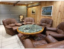 Wohnraum mit Holzwänden, einer Sitzgruppe aus braunen Ledersesseln und einem runden Tisch mit einer grünen Marmorplatte. Im Hintergrund ist eine Rolltür aus Metall sowie gerahmte Gemälde an der Holzwand zu sehen.