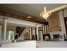 Eleganter Innenraum eines großen Wohnraums mit hohen Decken, verziert mit weißen Säulen mit goldenen Verzierungen, einer schmiedeeisernen Galerie, einem Kronleuchter und einer aufwendig gestalteten Kaminfassade. Helle Möbel und klassische Dekorelemente ergänzen das luxuriöse Ambiente.