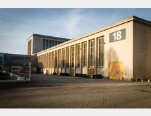 Großes, rechteckiges Gebäude mit beige-gelber Fassade und hohen Fenstern in symmetrischer Anordnung, nummeriert mit einer großen "18". Im Vordergrund ein gepflasterter Platz mit mehreren Müllcontainern und einem Parkplatzschild.