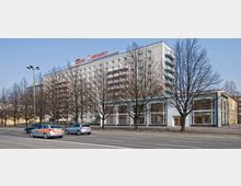 Mehrstöckiges Gebäude im Stil der Nachkriegsmoderne mit hellgrauer Fassade und großflächigen Fenstern im unteren Bereich, darüber ein Werbeschild. Vor dem Gebäude stehen kahle Bäume, und es verläuft eine mehrspurige Straße mit parkenden und fahrenden Autos.