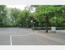 Kreuzung in einer verkehrsberuhigten Zone mit Fahrbahnmarkierungen, Verkehrsampeln und Verkehrsschildern für Radwege, umgeben von Bäumen und Grünflächen. Im Hintergrund sind Büsche und hohe Bäume zu sehen.