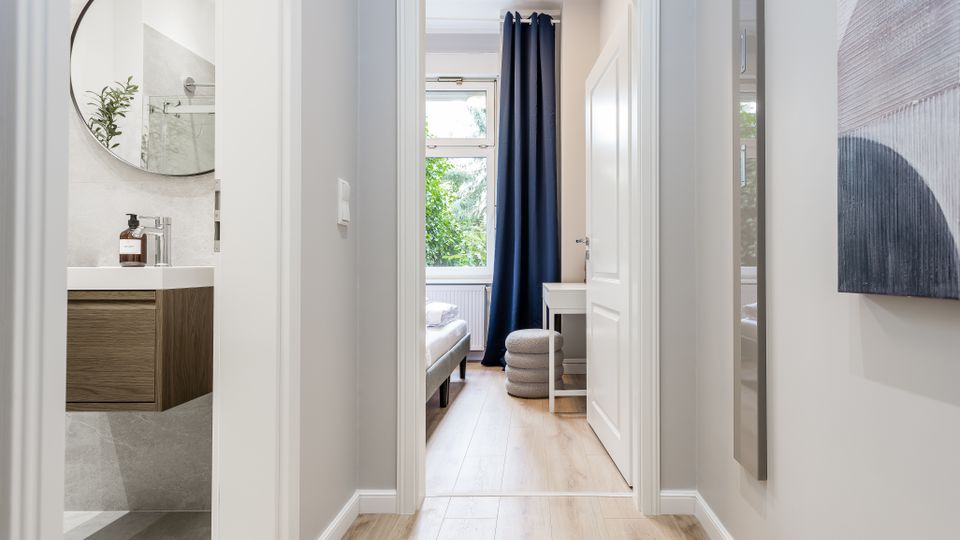 Heller, moderner Wohnungsflur mit hellen Holzdielen und weißen Türrahmen, Blick durch eine offene Tür in ein Schlafzimmer mit großem Fenster und dunkelblauem Vorhang. Links ist ein kleines Bad mit Waschtisch und rundem Spiegel zu sehen.