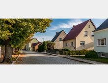 Eine gepflasterte Wohnstraße mit Einfamilienhäusern auf beiden Seiten, die überwiegend Satteldächer und helle Fassaden haben. Auf der linken Seite säumen große Bäume mit herbstlichem Laub den Gehweg, und die Umgebung wirkt ruhig und gepflegt.