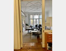 Blick in ein modernes Büro mit mehreren Schreibtischen und Stühlen, eingebettet in einen Raum mit hohen Decken und einer dekorativen Stuckverzierung. Im Hintergrund sind große Fenster und Regale mit Aktenordnern zu sehen, der Boden besteht aus Parkett und einem beigefarbenen Teppich.