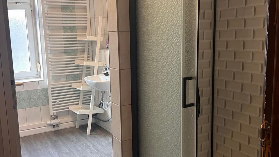 Badezimmer mit gefliester Dusche und mattierter Glas-Schiebetür, daneben eine strukturierte Wandverkleidung im Duschbereich. Im Hintergrund stehen ein Waschbecken, ein weißer Handtuchheizkörper und ein Fenster; der Boden ist in grauer Holzoptik gehalten.