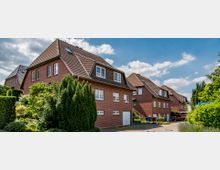 Straßenszene mit roten Backsteinhäusern in einer Wohnsiedlung; die Häuser haben steile Satteldächer mit roten Dachziegeln, mehrere Fenster und gepflegte Vorgärten mit Büschen und Bäumen. Der Himmel ist leicht bewölkt, und ein Straßenschild markiert die "Hagebuttenstraße".