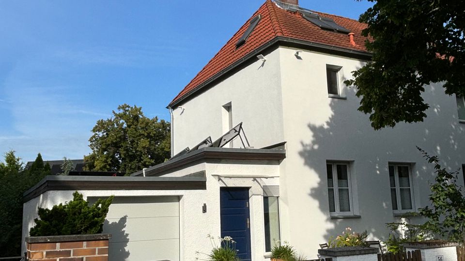 Einfamilienhaus mit weißer Fassade und rotem Ziegeldach, vorne mit blauem Eingang und angebauter Garage. Davor stehen ein niedriger Holzzaun sowie gemauerte Pfeiler, umgeben von Bäumen unter blauem Himmel.