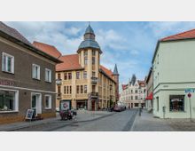 Straßenszene mit Kopfsteinpflaster in einem historischen Stadtkern; ein großes, gelbes Gebäude mit Turm und roten Dachziegeln steht im Mittelpunkt, umgeben von weiteren mehrstöckigen Häusern mit grünen und weißen Fassaden. Im Vordergrund befinden sich ein Laden und ein Bistro, mit vereinzelt parkenden Autos und Passanten.