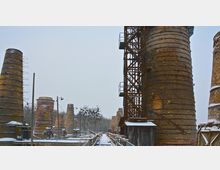 Industrielle Anlage mit mehreren hohen, runden Schornsteinen aus Backstein, teilweise mit metallischen Gerüsten umgeben, im Winter bei Schnee. Im Hintergrund sind Bäume sichtbar.