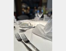 Ein festlich gedeckter Tisch mit weißer Tischdecke, glänzendem Besteck, gefaltetem Stoffservietten, Weingläsern und anderen Geschirrteilen im Vordergrund; der Hintergrund ist unscharf mit Lichtern und dunklen Möbeln.