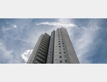 Hochwinkelansicht eines grauen Hochhauses mit Balkonen und Fenstern vor einem teilweise bewölkten, blauen Himmel. Das Gebäude hat eine geometrische, moderne Architektur und erhebt sich steil in die Höhe.