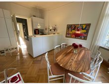 Helle Wohnküche mit Holzboden, einem ovalen Esstisch aus Holz mit weißen Stühlen und einem Blumengesteck als Tischdekoration. Im Hintergrund eine weiße Einbauküche mit hochmodernem Design, neben einem bunten Gemälde an der Wand; ein Fenster mit Vorhängen auf der rechten Seite.