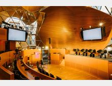 Moderner Konferenzraum mit geschwungenen Holzflächen und gläsernen Kuppelstrukturen. Der Raum verfügt über eine runde Sitzanordnung, eine Bühne mit schwarzen Stühlen, zwei große Bildschirme und technische Ausstattung wie Kameras und Beleuchtung.