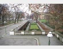 Parkanlage mit symmetrischen Rasenflächen und gepflegten Hecken, umgeben von Gehwegen und einer steinernen Balustrade. Im Hintergrund sind Wohngebäude zu sehen, und kahle Bäume säumen den Bereich, was auf eine Herbst- oder Winteratmosphäre hinweist.